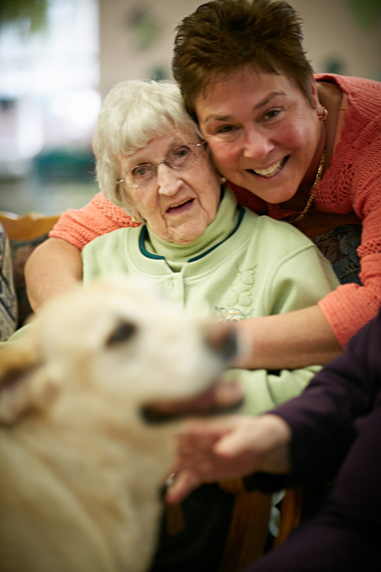 A caregiver hugs an elderly woman, with a dog in the foreground. A caregiver hugs an elderly woman, with a dog in the foreground.